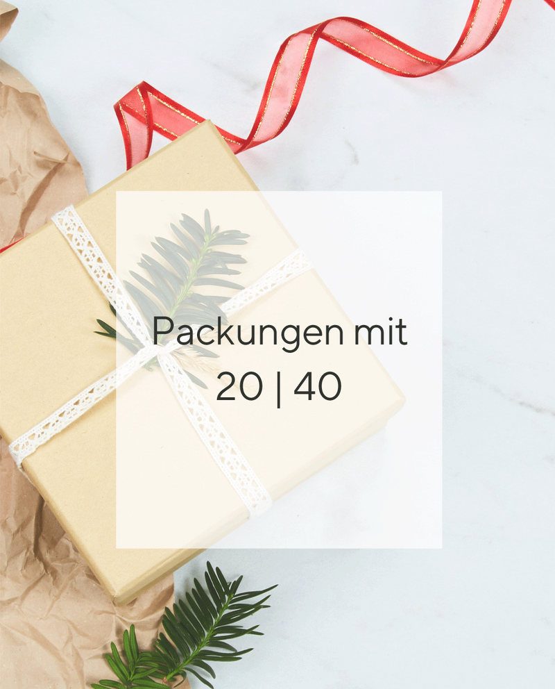 Ein tiefroter und ein dunkelgrüner ballonförmiger Aufkleber mit einem Weihnachtsbild und einem Weihnachtsgruss. Die Aufkleber sitzen auf einem in cremefarbigen eingepacktem Geschenk, ein Weihnachtsbaumzweig steckt in dem weißen umwickeltem Geschenkband. Drumherum liegt etwas braunes Paketpapier und rotes Geschenkband. Der Hintergrund ist hellgrauer Marmor.