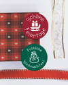 Eine rote karierte Serviette mit kleinen Weihnachtsbaummotiven, ein Stück weißes Geschenkband mit Goldborte und goldenen Aplikationen und ein rotes Geschenkband auf weißem Hintergrund. In der Mitte sind 2 Runde Weihnachtsaufkleber abgebildet. Der obere ist rot mit dem Text "Schöne Weihnachten" und einem Schlitten mit Geschenk als Motiv. Der untere Aufkleber ist Tannengrün, hat den Text " Fröhliche Weihnachten" und einen Schneemann als Motiv.