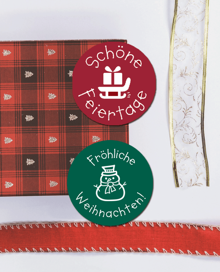 Eine rote karierte Serviette mit kleinen Weihnachtsbaummotiven, ein Stück weißes Geschenkband mit Goldborte und goldenen Aplikationen und ein rotes Geschenkband auf weißem Hintergrund. In der Mitte sind 2 Runde Weihnachtsaufkleber abgebildet. Der obere ist rot mit dem Text "Schöne Weihnachten" und einem Schlitten mit Geschenk als Motiv. Der untere Aufkleber ist Tannengrün, hat den Text " Fröhliche Weihnachten" und einen Schneemann als Motiv.