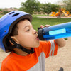 Dunkelhaariger Junge, trägt einen blauen Fahrradhelm und ein orange-grau gestreiftes T-Shirt. Seine Augen sind geschlossen und er trinkt aus einer blauen Wasserflasche. Die Wasserflasche ist mit einem hellblauen Aufkleber versehen. Der Aufkleber ist mit dem Namen William Wilson in der Schriftart School Print und einem Totenkopf als Motiv beschriftet. Im Hintergrund befindet sich ein Park und ein Kinderspielplatz.