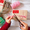 Auf einem Holztisch sind zwei Hände zu sehen, die gerade ein Weihnachtsgeschenk verpacken. Die rechte Hand schneidet mit einer Schere ein rot-weißes Weihnachtsband durch. Links oben klebt ein roter Weihnachtsgeschenkaufkleber, bedruckt mit drei Tannenbäume und einer Gruß der Familie Johnstons. Links unten vom Geschenk liegt ein anderes in grün- und links oben ein in beige eingewickeltes Geschenk. In der Mitte befindet sich eine Kordel mit dem rot-weißen Geschenkband.