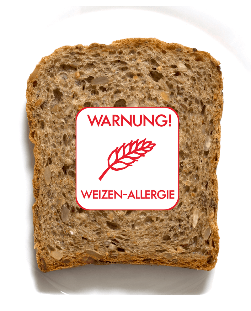 Eine Scheibe Vollkornbrot ist auf weißem Hintergrund abgebildet. Auf dem Vollkornbrot befindet sich ein weißer Allergie-Aufkleber mit einer Weizenähre und rotem Text "Warnung! Weizen-Allergie" in der Schriftart Clear und Simple abgedruckt.