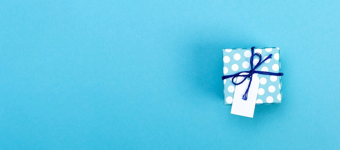 Ein Geschenk in hellblauen, weiß gepunkteten Geschenkpapier, mit marineblauer Schnur und einem weißen leeren Namensschild gebunden, welches auf einem schlichten blauen Hintergrund liegt