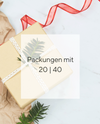 Ein in beigen Geschenkpapier eingepacktes Geschenk , verziert mit einem Weihnachtsbaumzweig, umwickelt mit cremefarbigen Geschenkband. Es liegen etwas braunes Packpapier und ein Stück rotes Geschenkband verstreut herum. Der Hintergrund ist hellgrauer Marmor.
