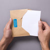 Die linke Hand hält einen braunen Briefumschlag, während die rechte Hand eine weiße Karte in den Umschlag einlegt. Ein blauer Adressaufkleber ist auf den Briefumschlag geklebt. 